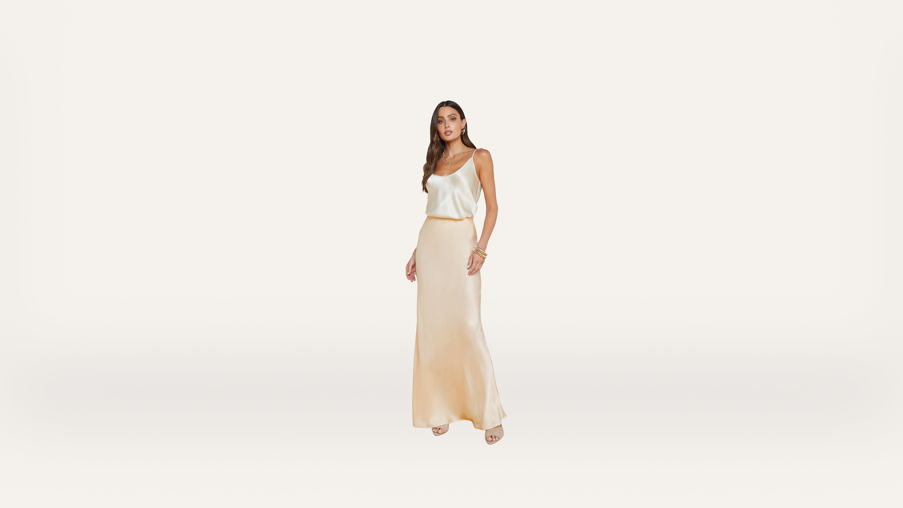 A woman stands against a plain white background, wearing a cream-colored satin tank top and a matching long skirt with beige heels. She has long brown hair and is accessorized with bracelets.