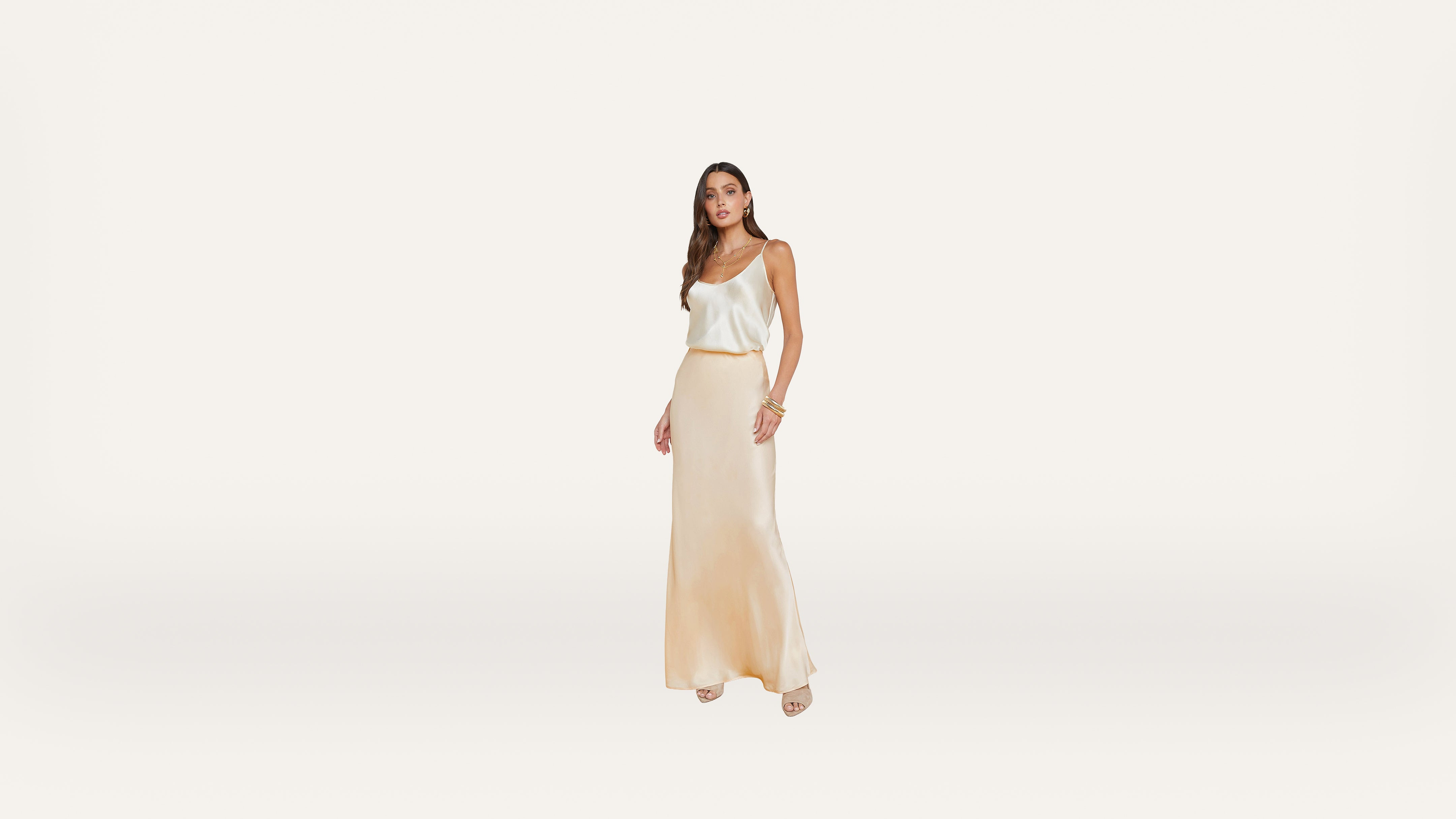 A woman stands against a plain white background, wearing a cream-colored satin tank top and a matching long skirt with beige heels. She has long brown hair and is accessorized with bracelets.