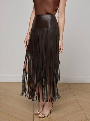 A person wears the L'AGENCE Karolina Fringe Skirt—a high-waisted vegan leather style that falls below the knees—paired with a sleeveless brown top and beige open-toe heels, standing on a wooden floor against a light background.