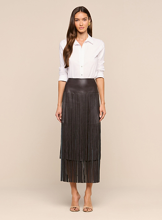 A woman stands facing forward, wearing a white button-down shirt tucked into the high-waisted Karolina Fringe Skirt, crafted from black vegan leather with long, dramatic fringe. She completes the look with beige open-toe heels against a plain, neutral background.