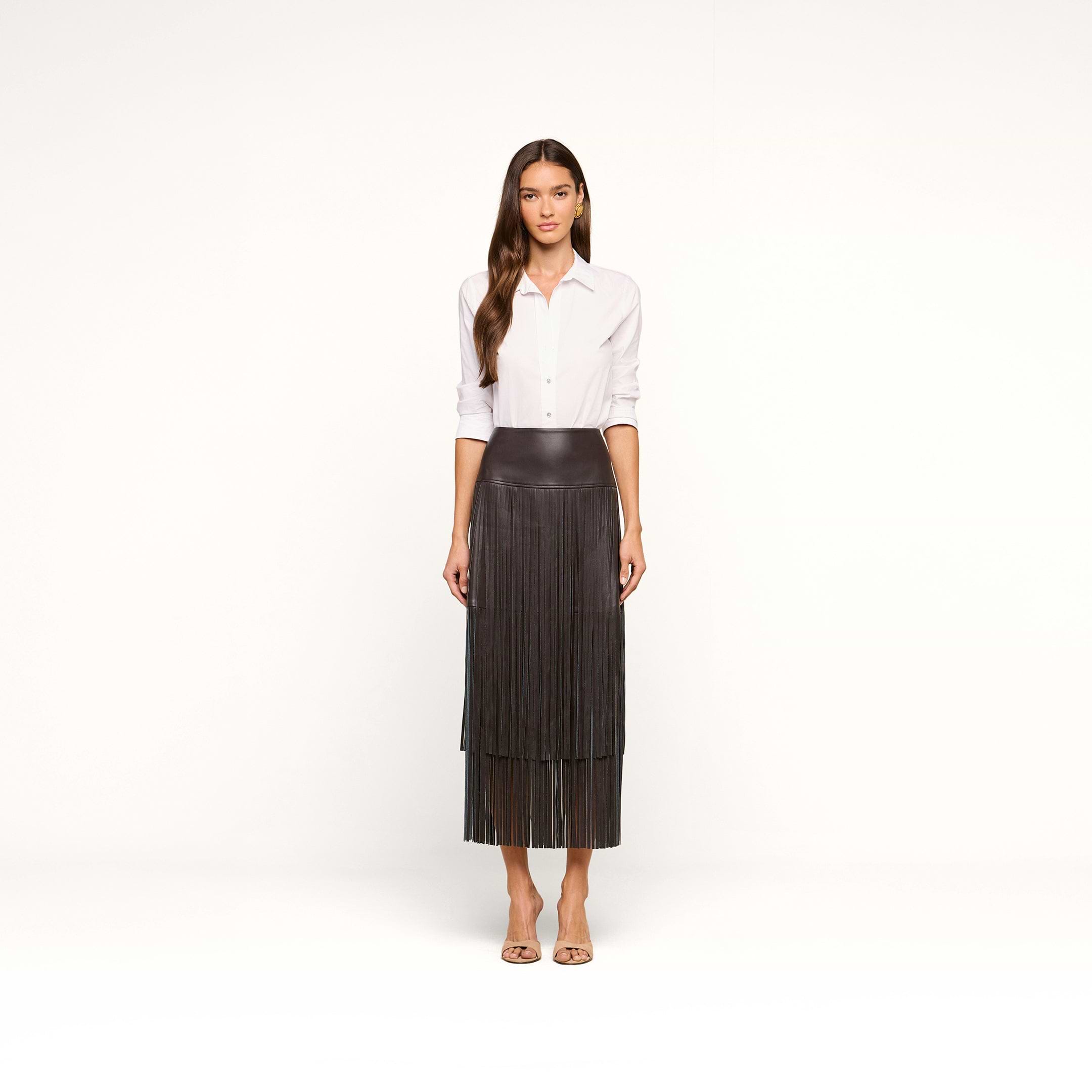 A woman stands facing forward against a plain white background, wearing a white button-down shirt, the Karolina Fringe Skirt in black, and beige open-toed heels.