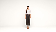 A woman stands against a plain white background wearing a white button-up shirt, a long dark pleated skirt, and open-toed sandals. She has long hair and a neutral expression.