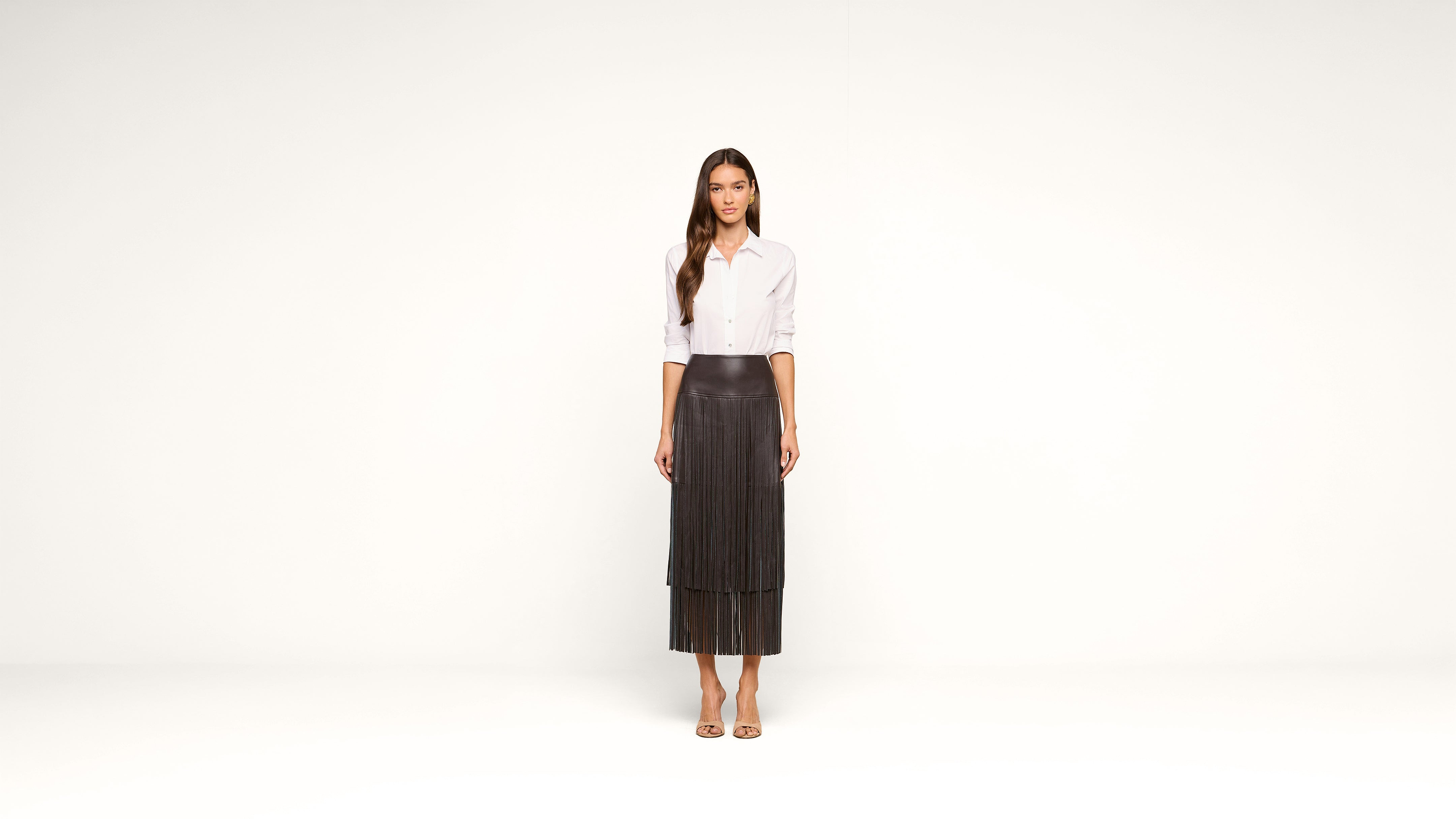 A woman stands against a plain white background wearing a white button-up shirt, a long dark pleated skirt, and open-toed sandals. She has long hair and a neutral expression.