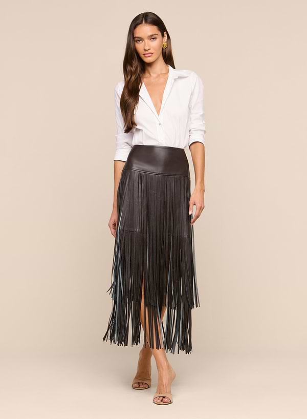 A woman stands facing forward on a plain white background, wearing a white blouse, neutral heeled sandals, and the Karolina Fringe Skirt in black vegan leather.