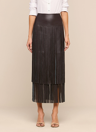 A woman stands against a beige background wearing the Karolina Fringe Skirt—a black high-waisted skirt with two layers of long fringe—paired with a white button-up shirt and open-toed nude heels.