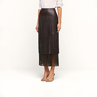 Wearing the Karolina Fringe Skirt, a high-waisted black vegan leather piece with fringe details, along with a white long-sleeve shirt and open-toe heels, a woman stands against a plain white background.