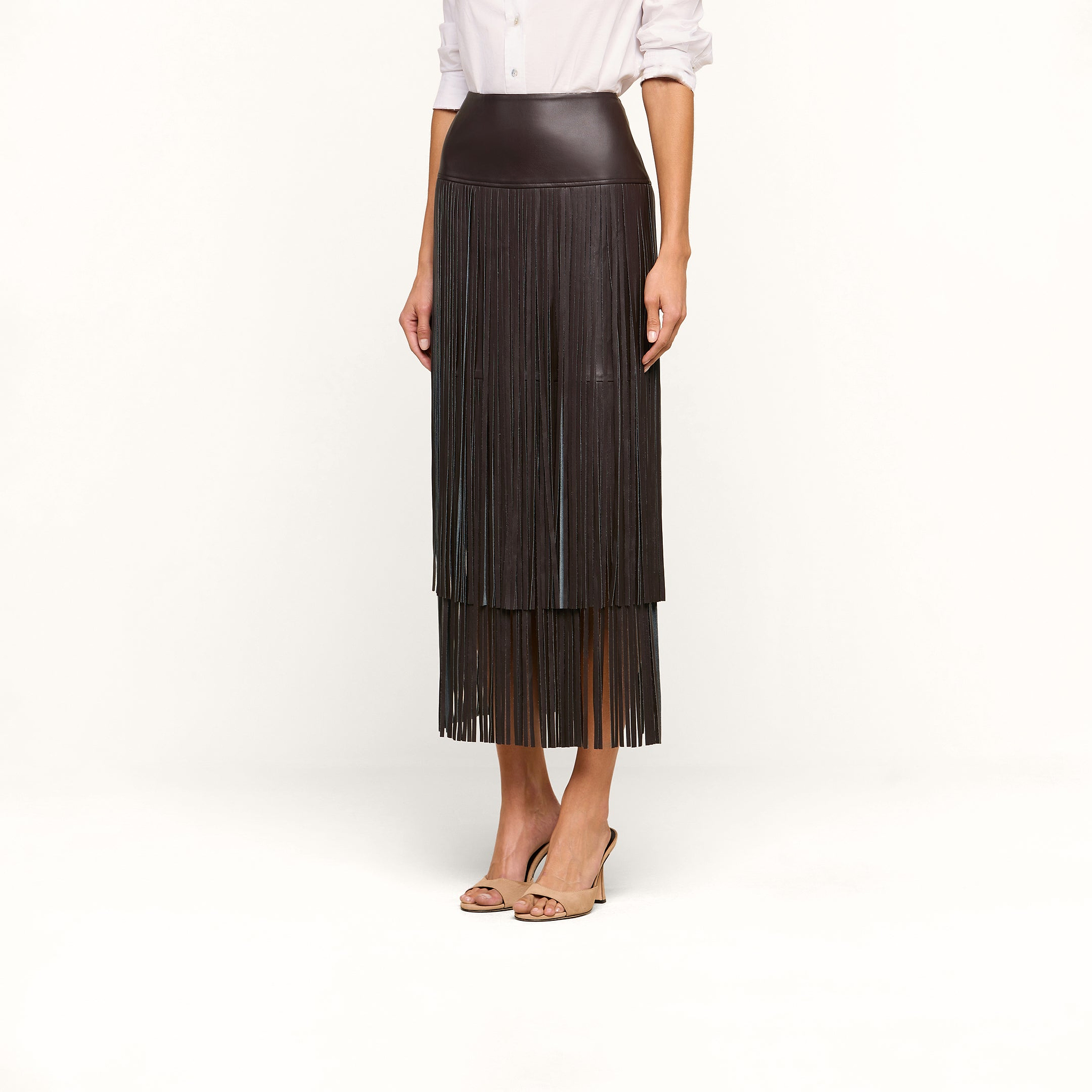Wearing the Karolina Fringe Skirt, a high-waisted black vegan leather piece with fringe details, along with a white long-sleeve shirt and open-toe heels, a woman stands against a plain white background.