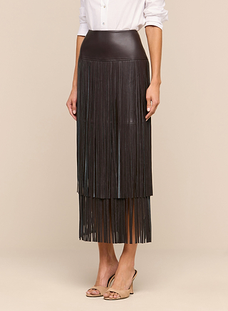 A person wears the Karolina Fringe Skirt, a black high-waisted vegan leather skirt featuring long fringe details, paired with a white long-sleeve shirt and nude open-toe heels, standing against a plain beige background.