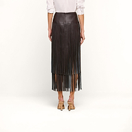 A person wearing a white long-sleeve shirt, the Karolina Fringe Skirt in black vegan leather, and beige heeled shoes stands with their back to the camera against a plain white background.