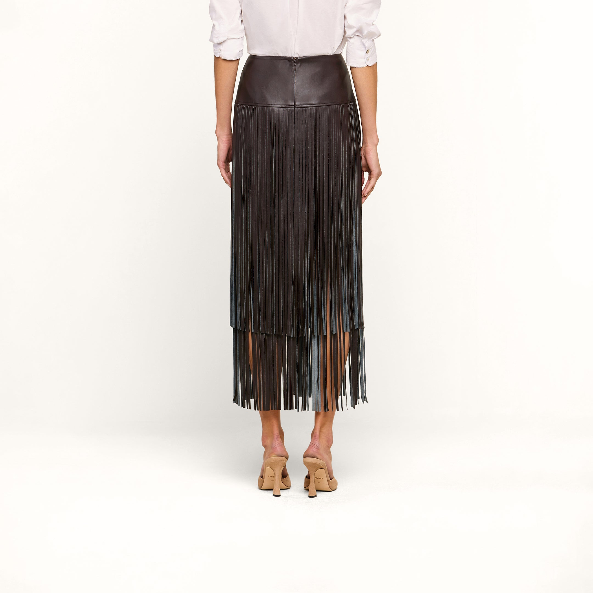 A person wearing a white long-sleeve shirt, the Karolina Fringe Skirt in black vegan leather, and beige heeled shoes stands with their back to the camera against a plain white background.