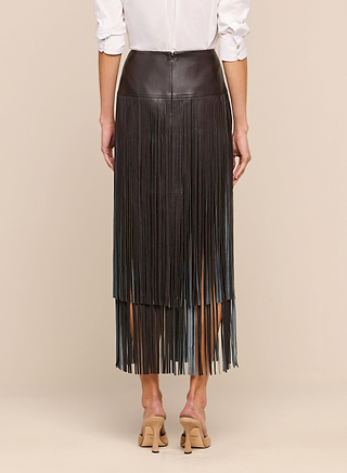 A person stands facing away against a neutral background, wearing the Karolina Fringe Skirt in brown vegan leather with a white shirt and beige heeled sandals.