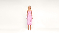 A woman stands facing forward wearing a light pink, sleeveless, knee-length satin slip dress and open-toe heels against a plain white background.