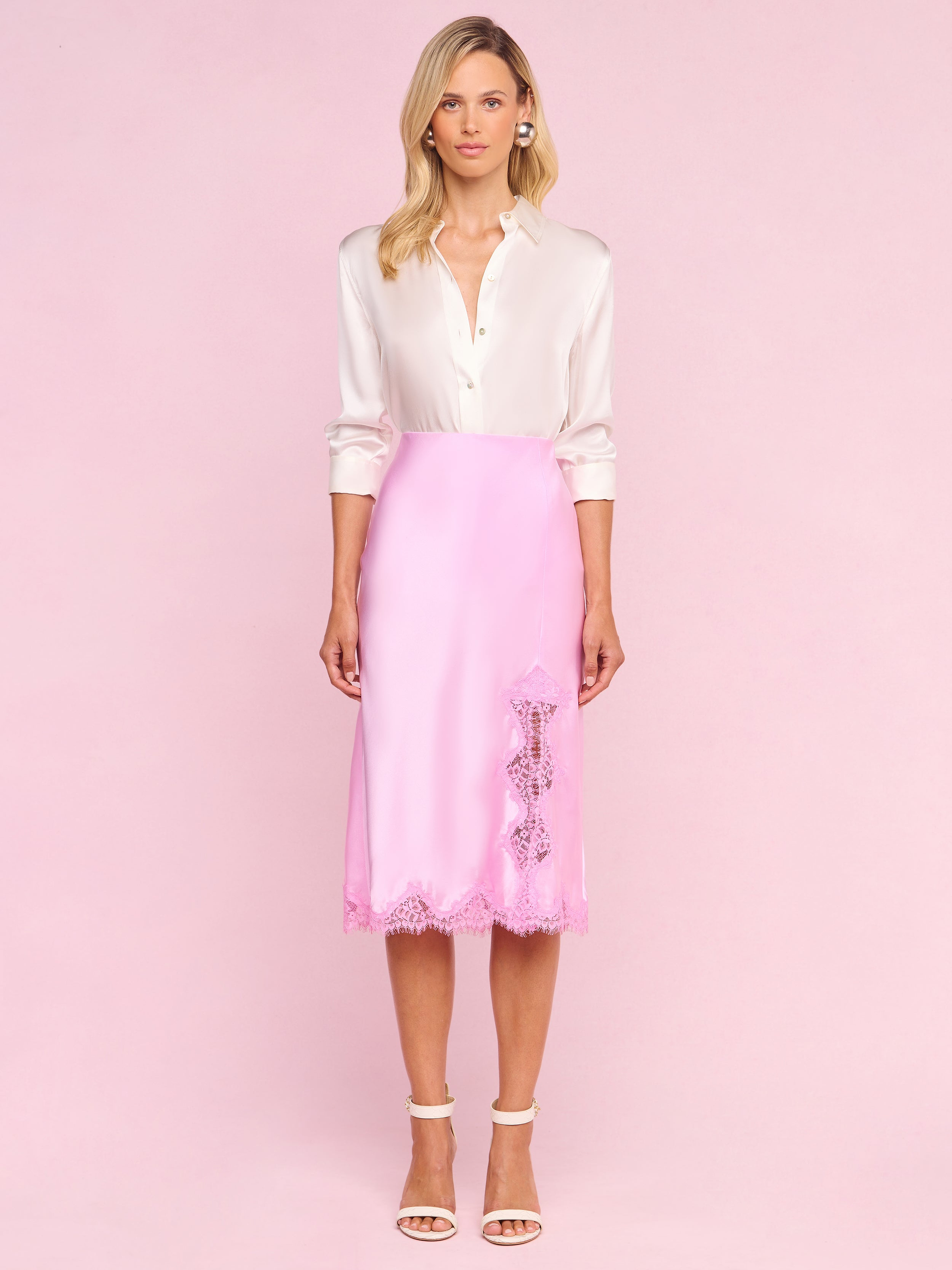 A woman stands facing forward, wearing a white long-sleeve blouse tucked into the Tico Silk Lace Skirt—a pink silk midi skirt featuring eyelash lace details—and white high-heeled sandals, evoking a Legally Blonde-inspired style against a plain white background.