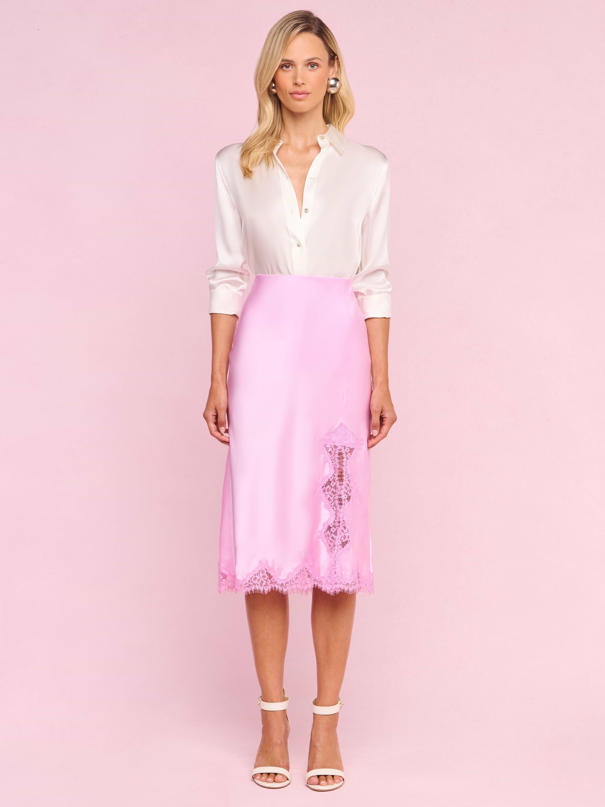 A woman stands facing forward, wearing a white long-sleeve blouse tucked into the Tico Silk Lace Skirt—a pink silk midi skirt featuring eyelash lace details—and white high-heeled sandals, evoking a Legally Blonde-inspired style against a plain white background.
