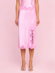Wearing the Tico Silk Lace Skirt—a shiny pink silk midi skirt featuring eyelash lace trim and a front slit—a person stands in clear high-heeled sandals against a plain light background.