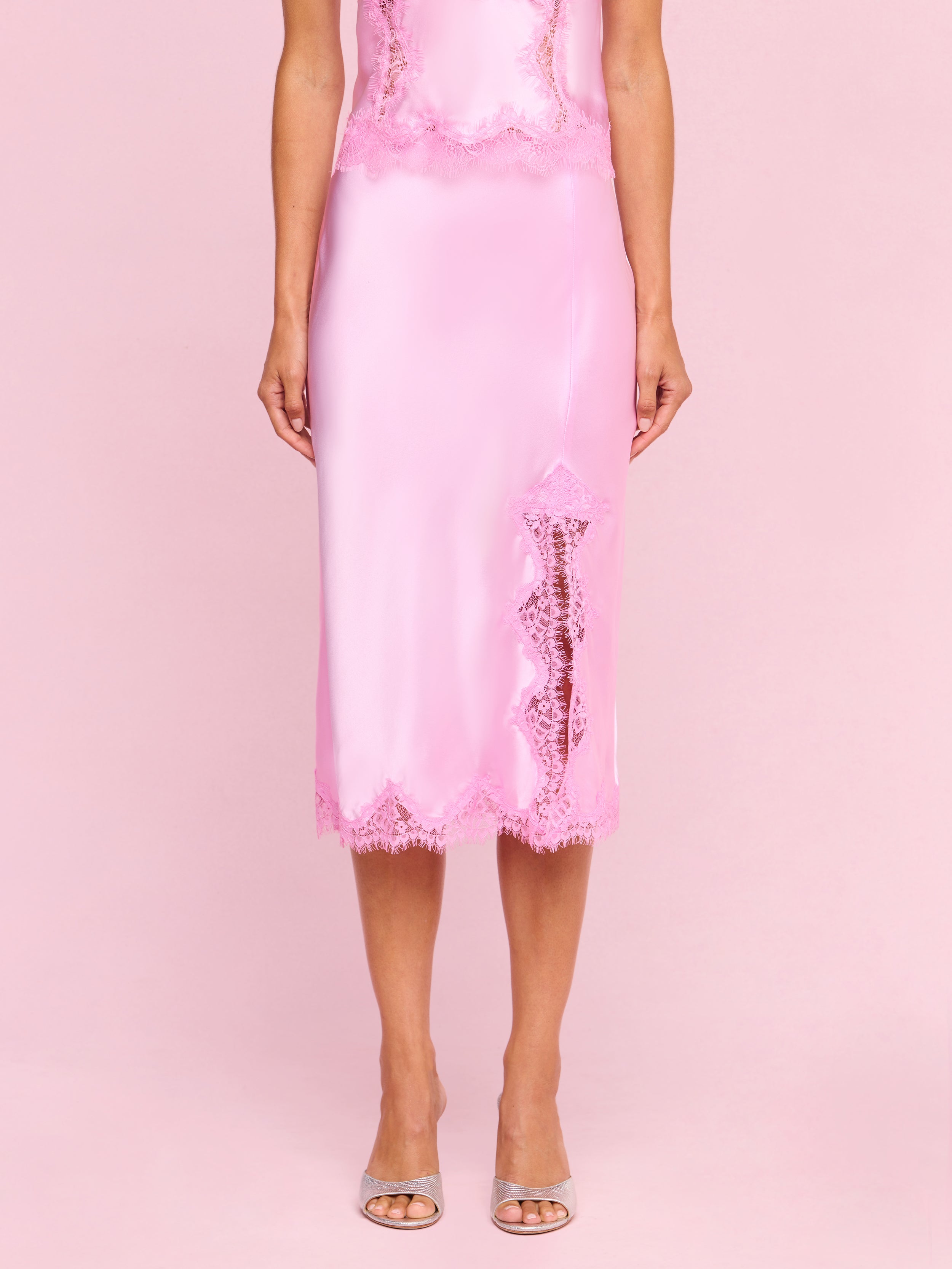 Wearing the Tico Silk Lace Skirt—a shiny pink silk midi skirt featuring eyelash lace trim and a front slit—a person stands in clear high-heeled sandals against a plain light background.