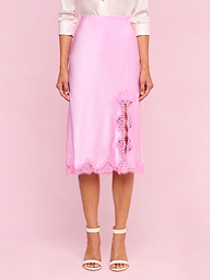 A woman wears the Tico Silk Lace Skirt—a shiny light pink silk midi skirt featuring eyelash lace trim and a lace detail on one side—paired with a light-colored blouse and white high-heeled sandals.