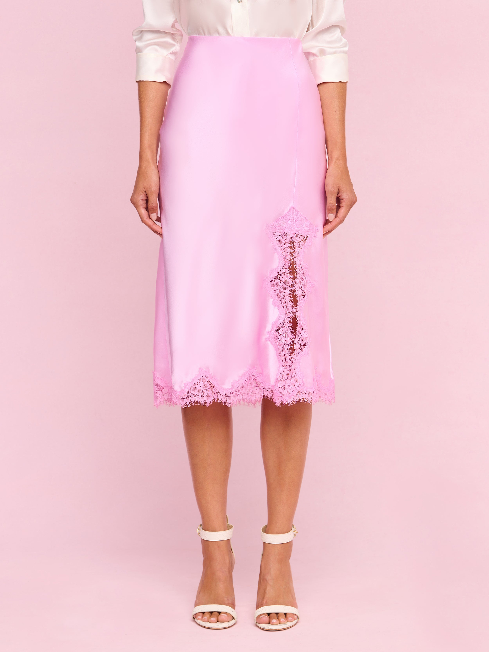 A woman wears the Tico Silk Lace Skirt—a shiny light pink silk midi skirt featuring eyelash lace trim and a lace detail on one side—paired with a light-colored blouse and white high-heeled sandals.