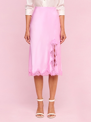 A woman stands against a pink background wearing the Tico Silk Lace Skirt from the Legally Blonde collection, paired with a white blouse and white high-heeled sandals.