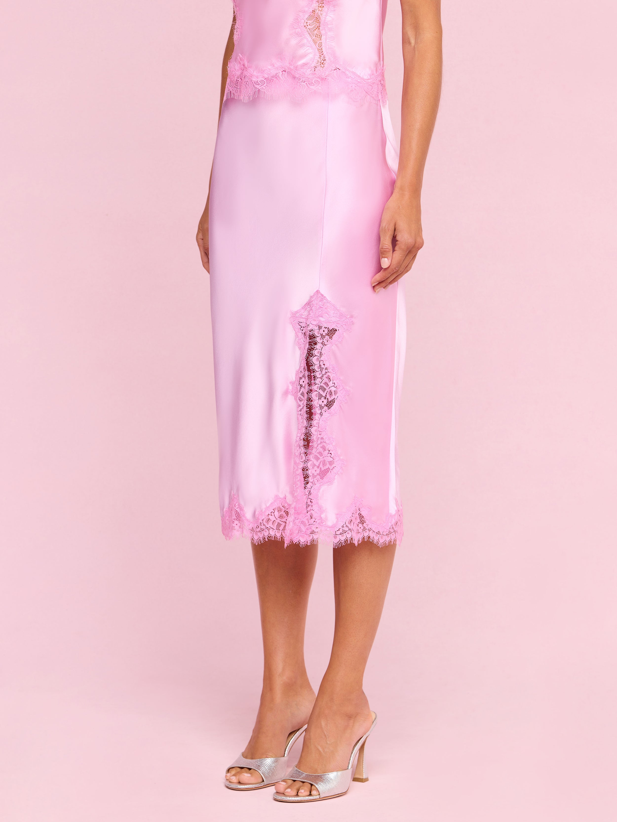 A woman wearing the Tico Silk Lace Skirt in light pink, styled with a coordinating lace dress featuring eyelash lace detailing and matching open-toe heeled sandals, stands against a plain off-white background.