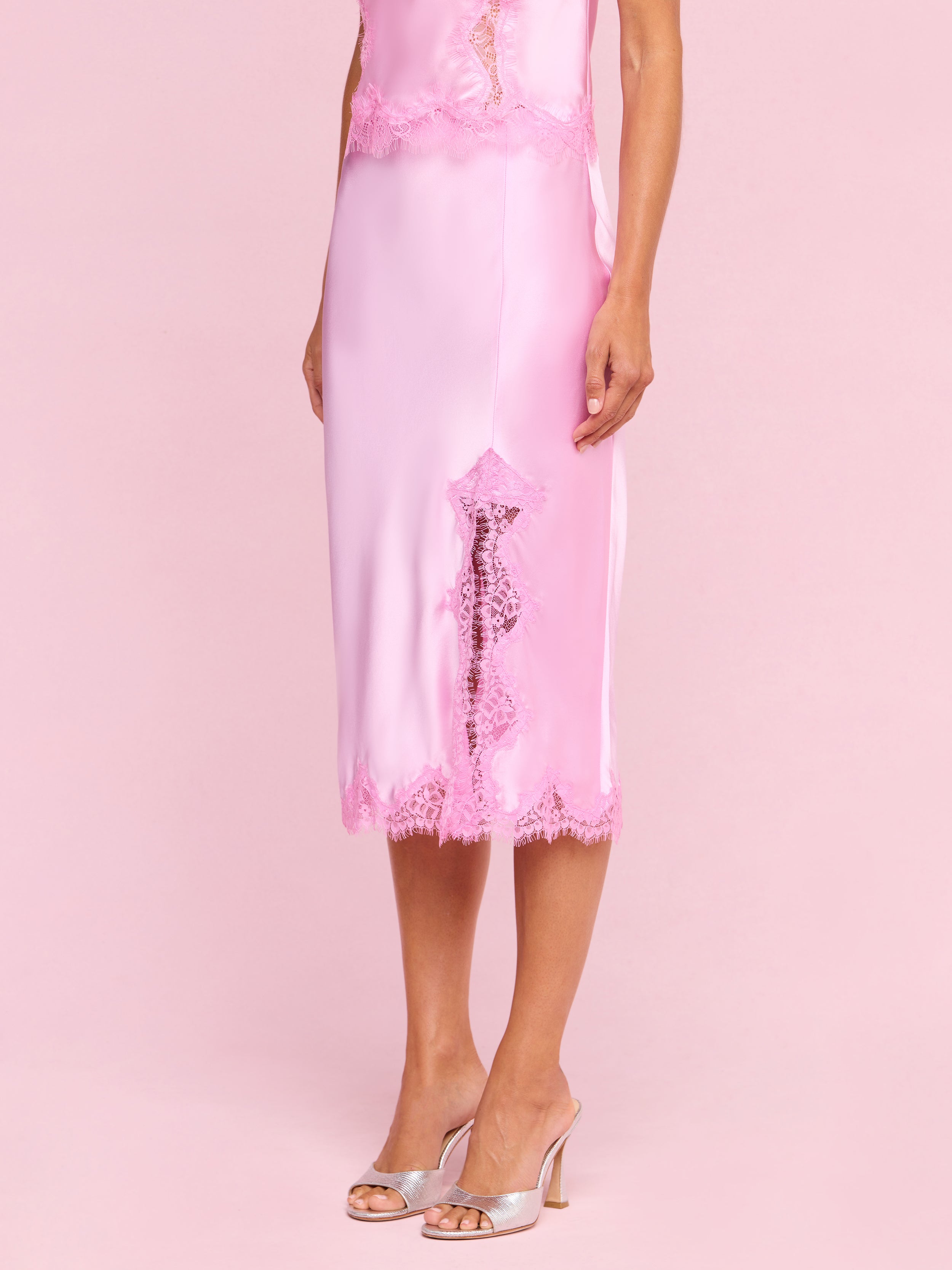 A woman wearing the Tico Silk Lace Skirt in light pink, styled with a coordinating lace dress featuring eyelash lace detailing and matching open-toe heeled sandals, stands against a plain off-white background.
