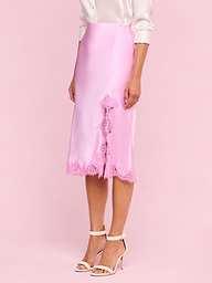 A person wearing the Tico Silk Lace Skirt—a silky pink midi skirt with eyelash lace trim—paired with a pale pink blouse with rolled sleeves and white high-heeled sandals stands against a plain light background.