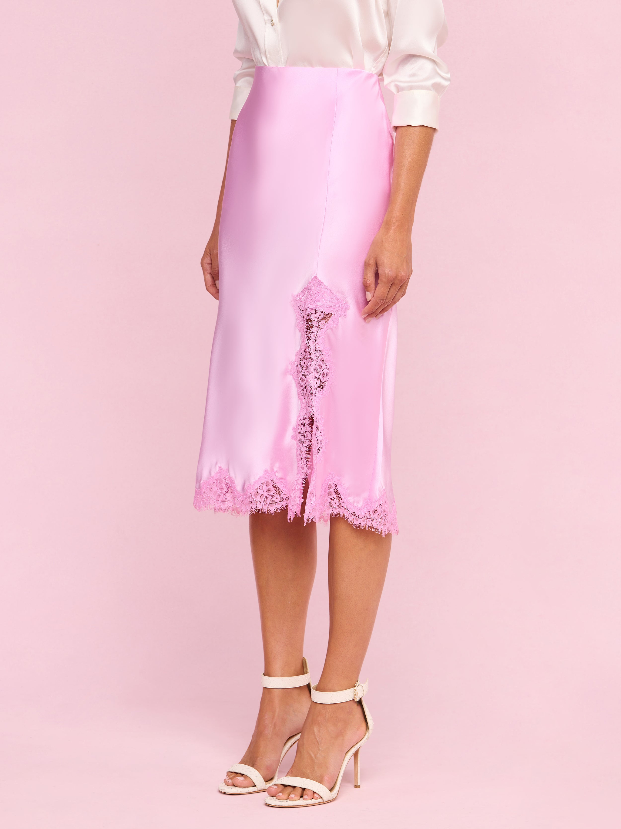 A person wearing the Tico Silk Lace Skirt—a silky pink midi skirt with eyelash lace trim—paired with a pale pink blouse with rolled sleeves and white high-heeled sandals stands against a plain light background.