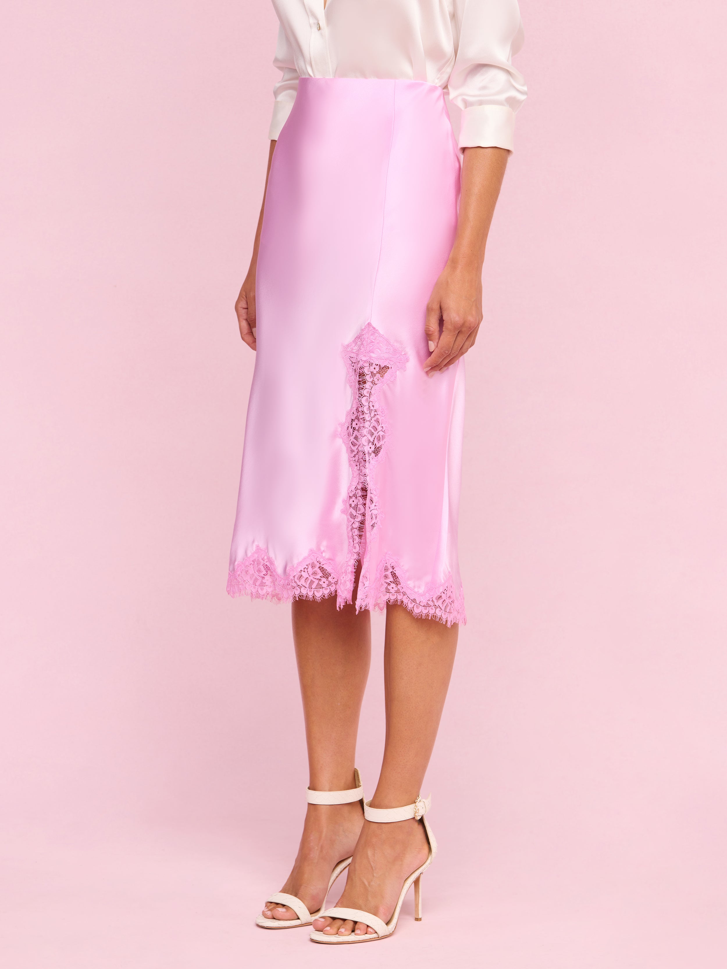 A person wearing the Tico Silk Lace Skirt—a silky pink midi skirt with eyelash lace trim—paired with a pale pink blouse with rolled sleeves and white high-heeled sandals stands against a plain light background.