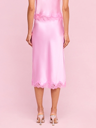 A woman stands facing away, wearing the Tico Silk Lace Skirt—a shiny pink silk midi skirt with eyelash lace trim—paired with a matching top and light pink high heels against a plain white background.