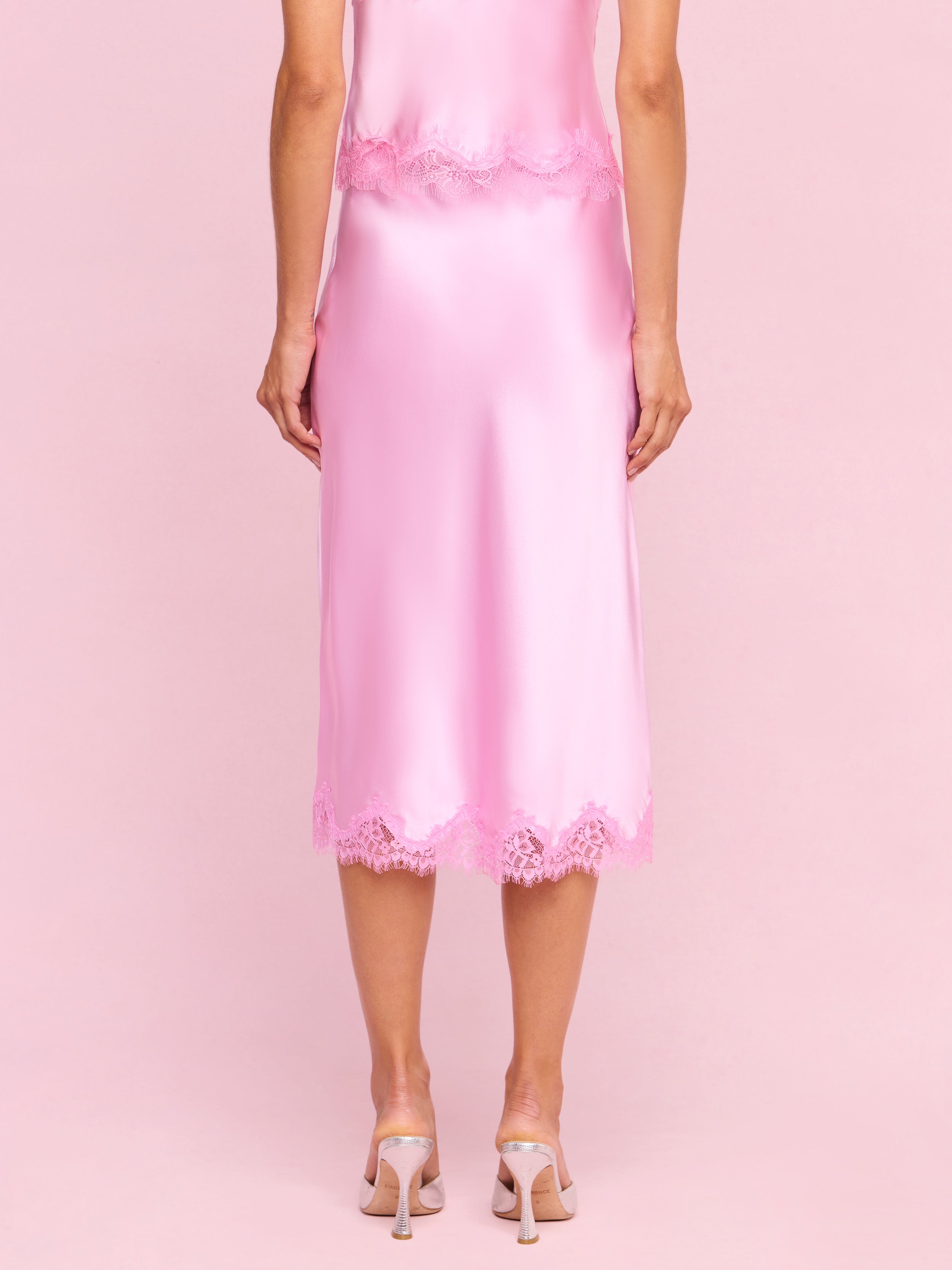 A woman stands facing away, wearing the Tico Silk Lace Skirt—a shiny pink silk midi skirt with eyelash lace trim—paired with a matching top and light pink high heels against a plain white background.