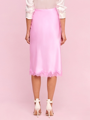 A person stands facing away, wearing the shiny pink Tico Silk Lace Skirt from the Legally Blonde collection with delicate trim and a cream blouse, paired with white high-heeled sandals against a pink background.