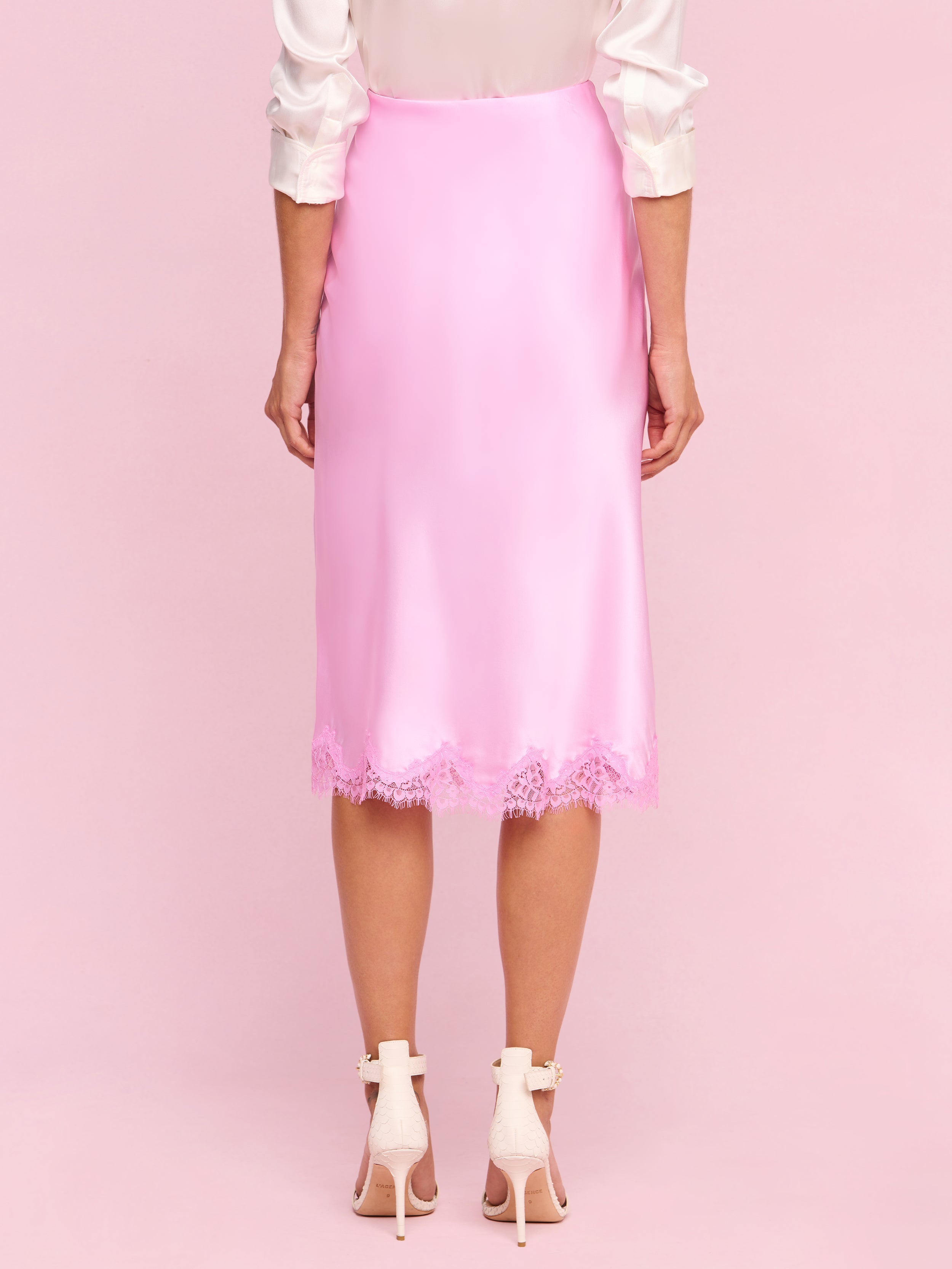 A person stands facing away, wearing the Tico Silk Lace Skirt in light pink, featuring delicate eyelash lace trim. The outfit is paired with a white blouse and light pink high-heeled shoes against a plain background, channeling classic Legally Blonde charm.