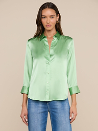 Una donna, con lo sguardo rivolto in avanti, indossa una camicetta Dani Silk verde chiaro con maniche a tre quarti e jeans blu, su uno sfondo beige.
