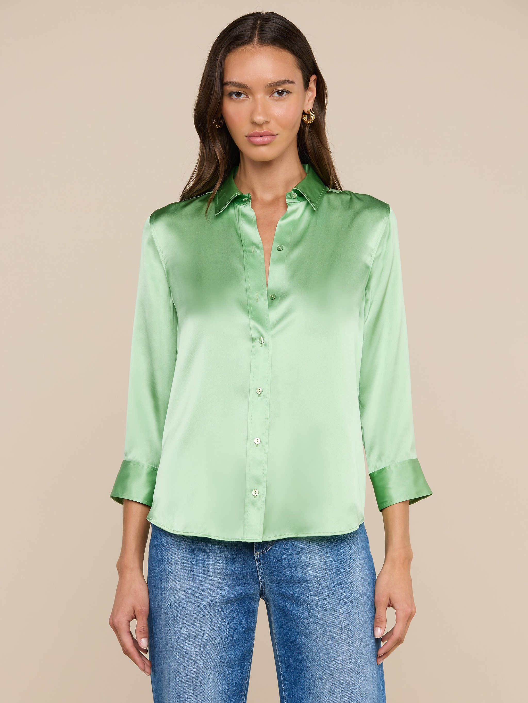 Una donna, con lo sguardo rivolto in avanti, indossa una camicetta Dani Silk verde chiaro con maniche a tre quarti e jeans blu, su uno sfondo beige.