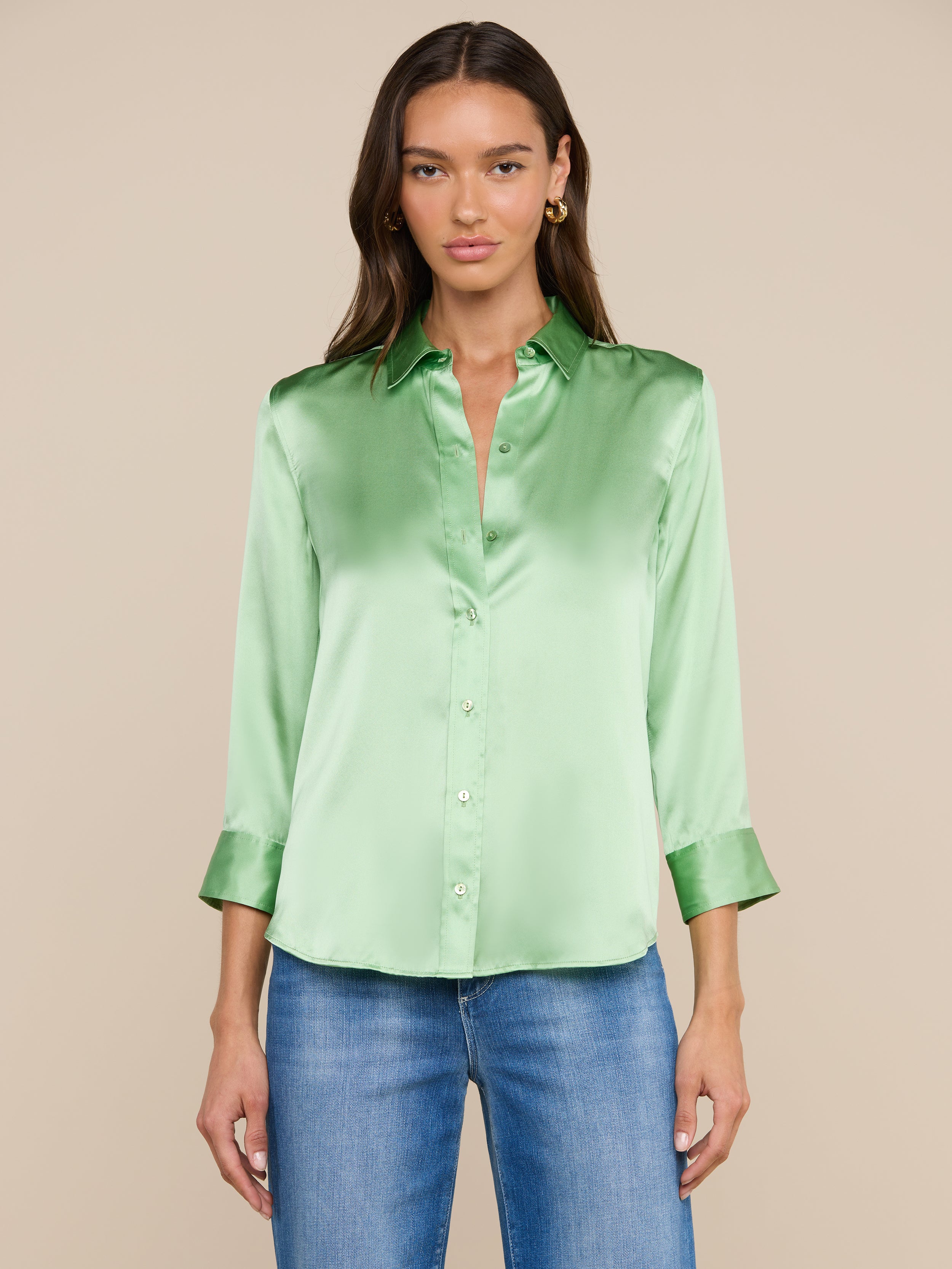 Una donna, con lo sguardo rivolto in avanti, indossa una camicetta Dani Silk verde chiaro con maniche a tre quarti e jeans blu, su uno sfondo beige.