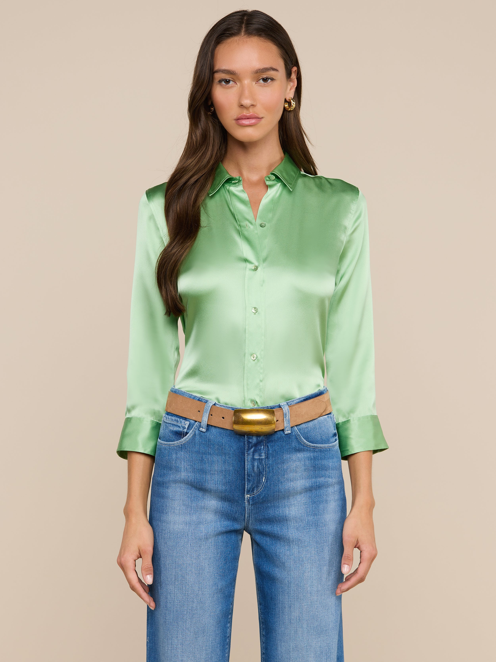 Una donna dai lunghi capelli castani indossa la camicetta Dani Silk — una camicetta di seta verde chiaro con bottoni e maniche a tre quarti — abbinata a jeans blu, una cintura color cuoio e orecchini a cerchio dorati, in piedi su uno sfondo beige.