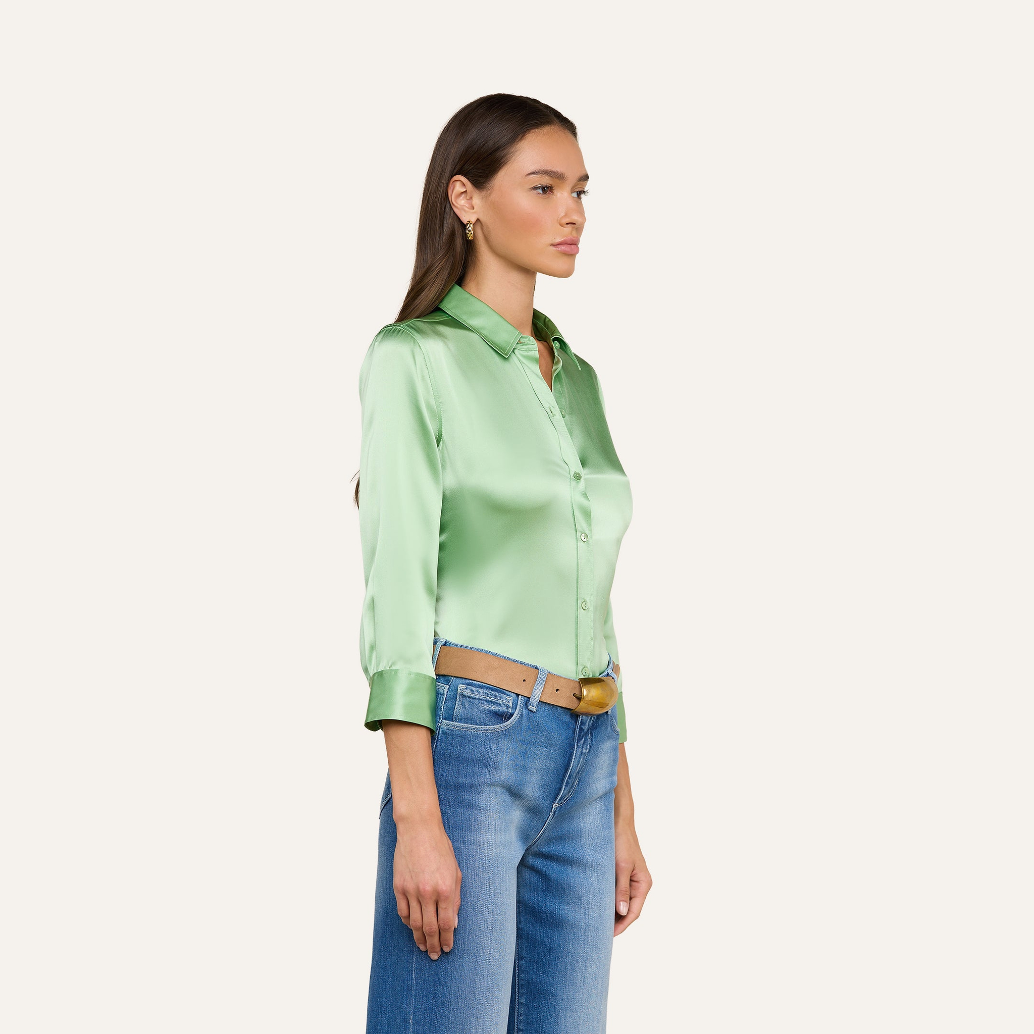 Una donna è in piedi di profilo su uno sfondo beige a tinta unita e indossa una camicetta Dani Silk verde chiaro con maniche a tre quarti, jeans blu e una cintura color cuoio.