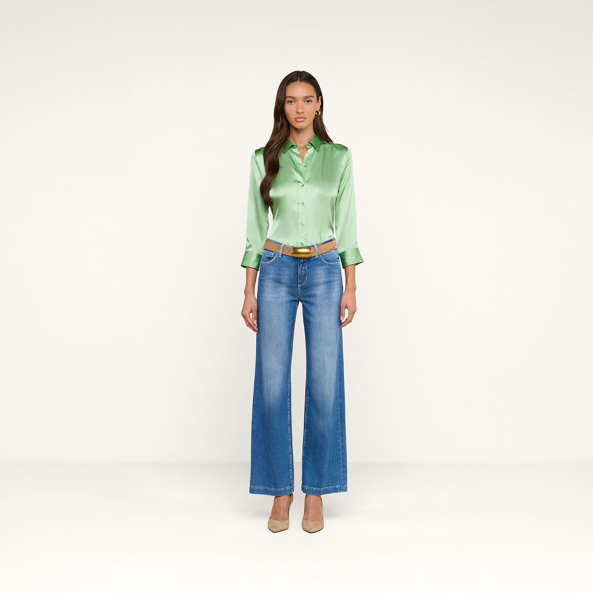 Una donna è ritratta di fronte con indosso una camicetta Dani Silk verde chiaro con maniche a tre quarti, jeans blu a vita alta e a gamba larga, una cintura marrone e tacchi beige, il tutto su uno sfondo beige a tinta unita.