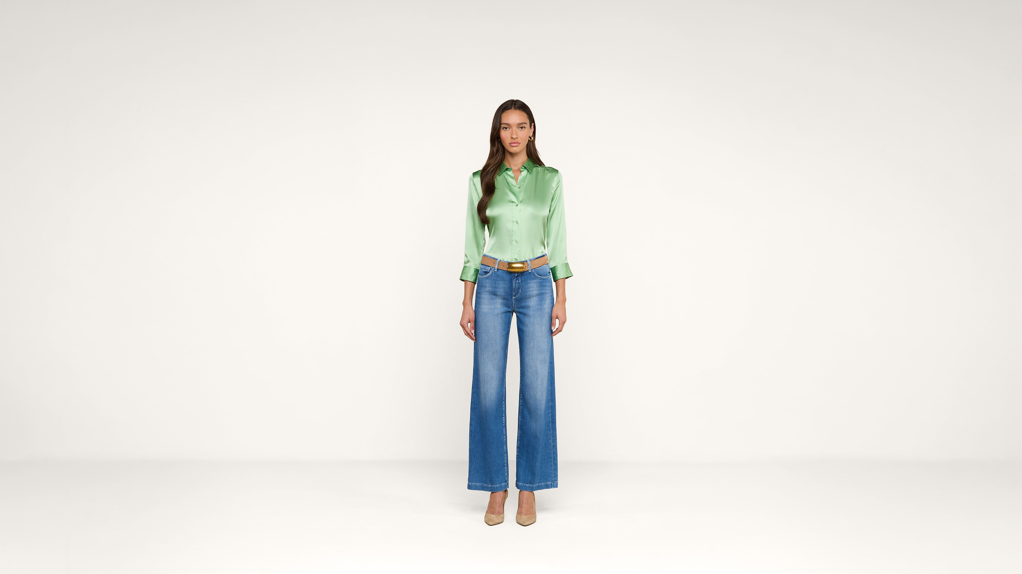 A woman stands facing forward, wearing a shiny green blouse, blue high-waisted wide-leg jeans, and beige heels, set against a plain white background.