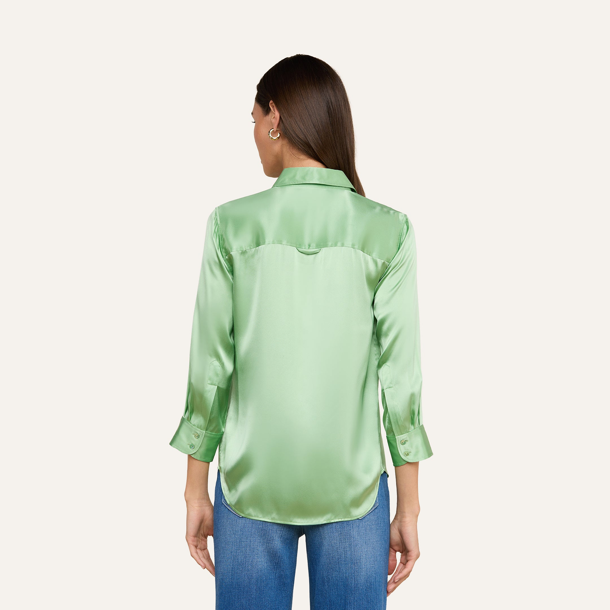 Si vede di spalle una donna dai lunghi capelli castani che indossa una camicetta Dani Silk verde chiaro lucida con maniche a tre quarti e jeans blu, su uno sfondo beige.