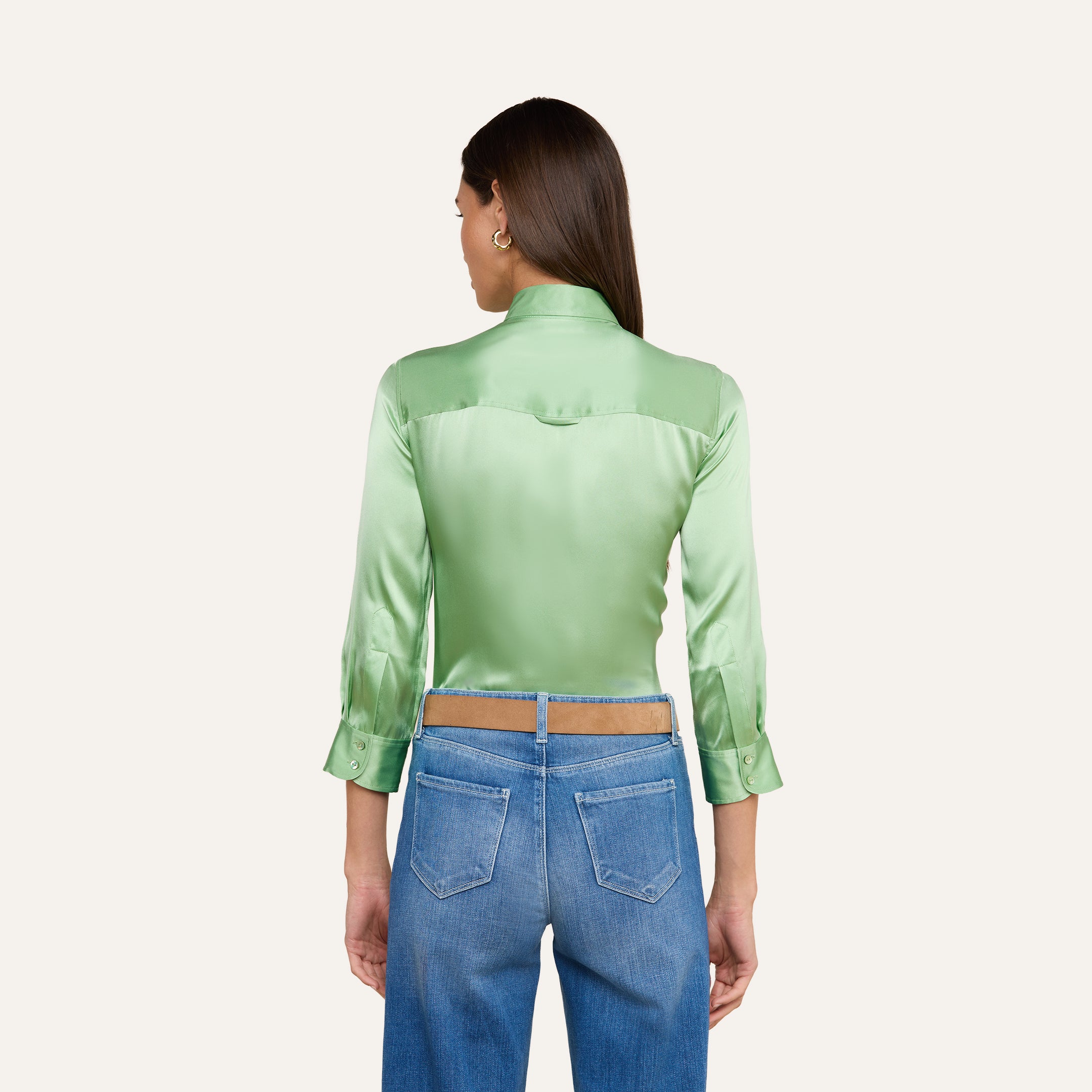 Si vede di spalle una donna dai lunghi capelli castani, che indossa una camicetta Dani Silk verde chiaro lucida con maniche a tre quarti e jeans blu a vita alta, in piedi su uno sfondo beige.