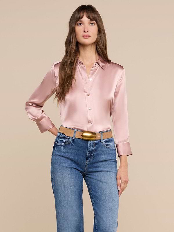 Una donna dai lunghi capelli castani indossa la camicetta Dani Silk, un modello lucido rosa chiaro abbottonato, infilata nei jeans blu e con una cintura color cuoio, in piedi davanti a uno sfondo beige a tinta unita.