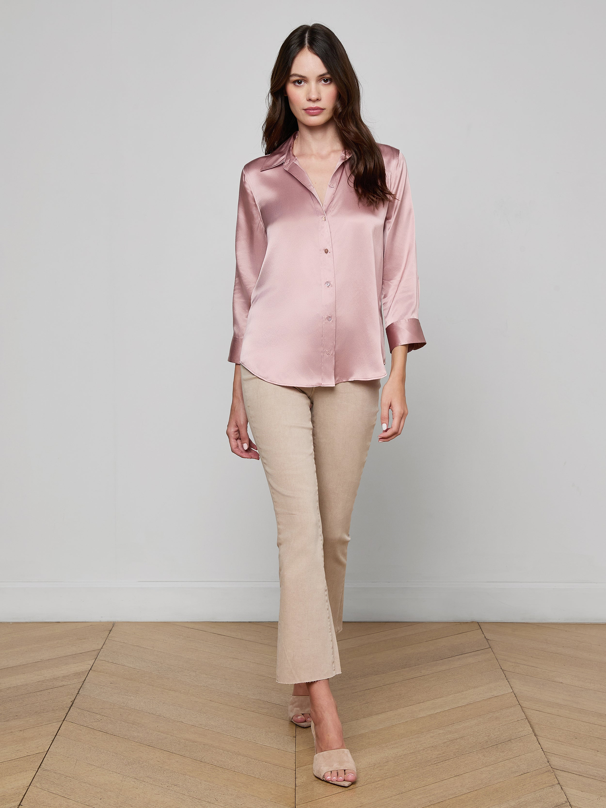 A woman wearing sunglasses, a light pink Dani Silk Blouse, blue high-waisted jeans, a brown belt, and brown heels stands against a plain white background, posing with one hand in her pocket.