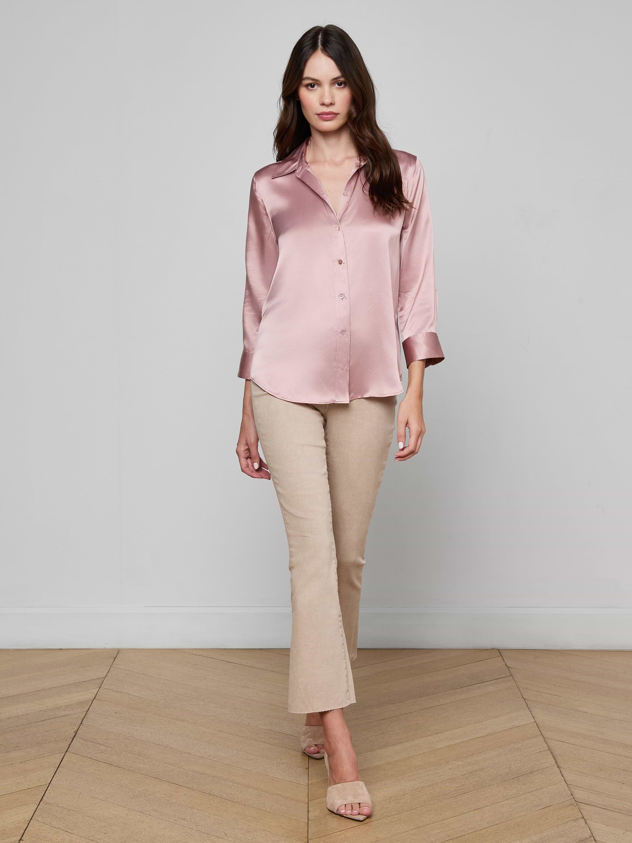 A woman wearing sunglasses, a light pink Dani Silk Blouse, blue high-waisted jeans, a brown belt, and brown heels stands against a plain white background, posing with one hand in her pocket.