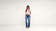 A woman wearing sunglasses, a pink blouse, blue wide-leg jeans, and brown heels stands against a plain white background.