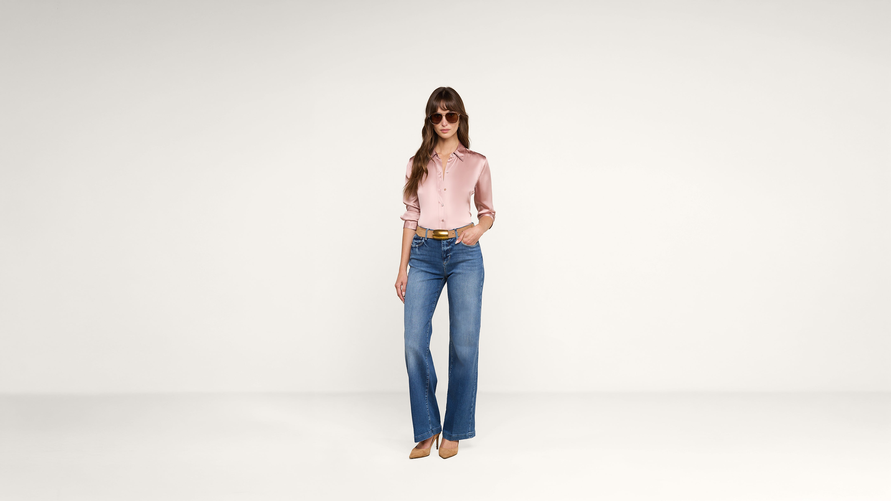 A woman wearing sunglasses, a pink blouse, blue wide-leg jeans, and brown heels stands against a plain white background.