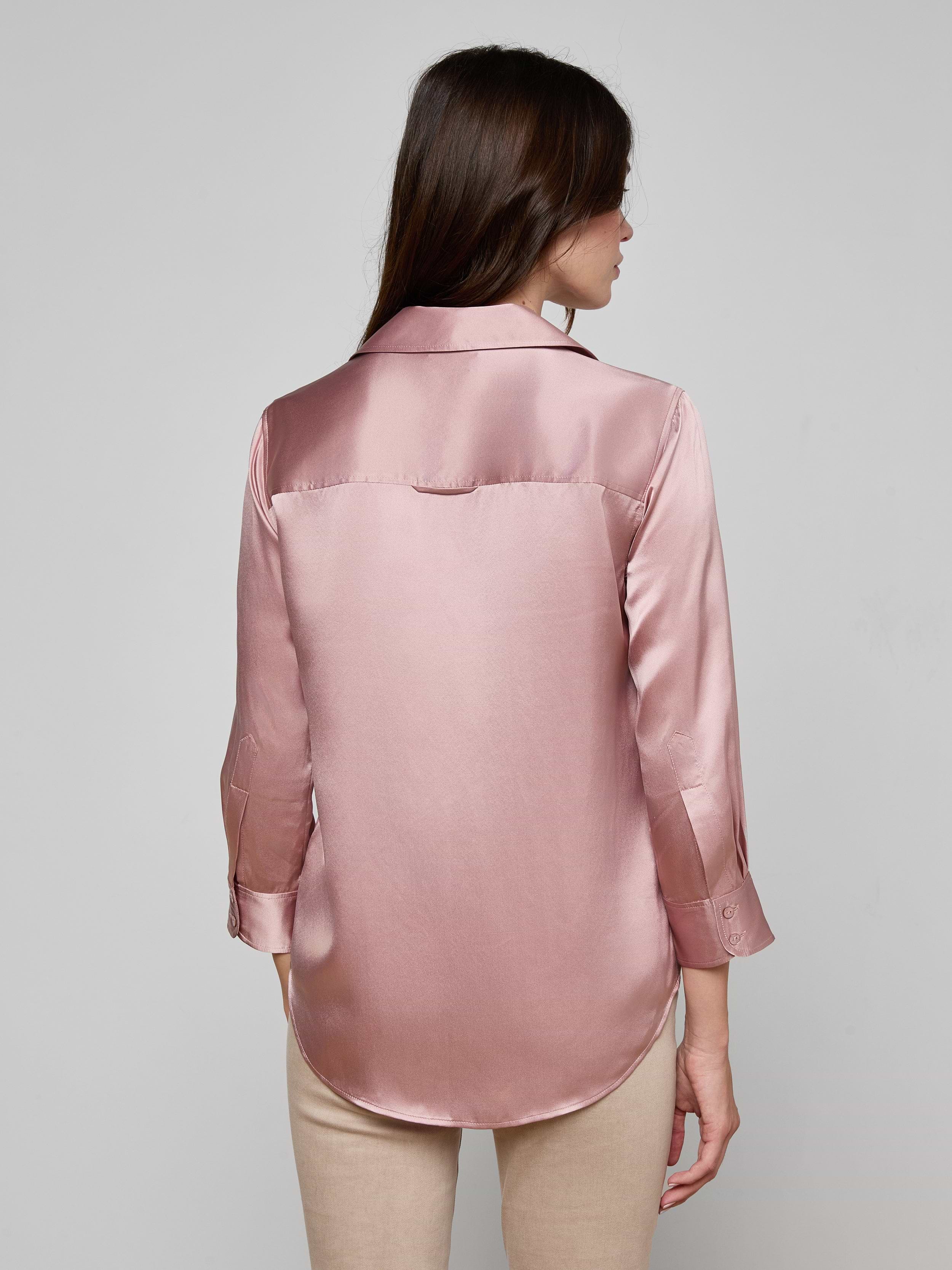 A woman stands with her back to the camera, wearing a Dani Silk Blouse in light pink, crafted from 100 silk charmeuse with long sleeves, paired with blue jeans, set against a plain white background.