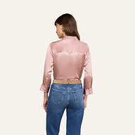 Una donna dai lunghi capelli castani e con orecchini doro è in piedi su uno sfondo beige; indossa la camicetta Dani Silk Blouse in rosa pallido a maniche lunghe e jeans blu.