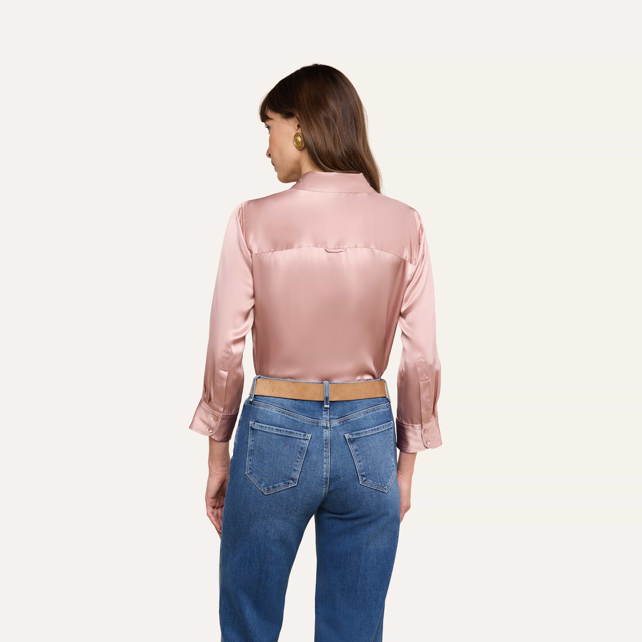 Una donna dai lunghi capelli castani e con orecchini doro è in piedi su uno sfondo beige; indossa la camicetta Dani Silk Blouse in rosa pallido a maniche lunghe e jeans blu.