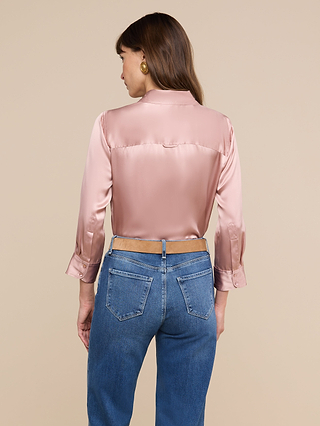 Une femme aux longs cheveux châtains et aux boucles doreilles dorées se tient debout devant un fond beige. Elle porte le chemisier « Dani Silk » rose pâle à manches longues et un jean bleu.